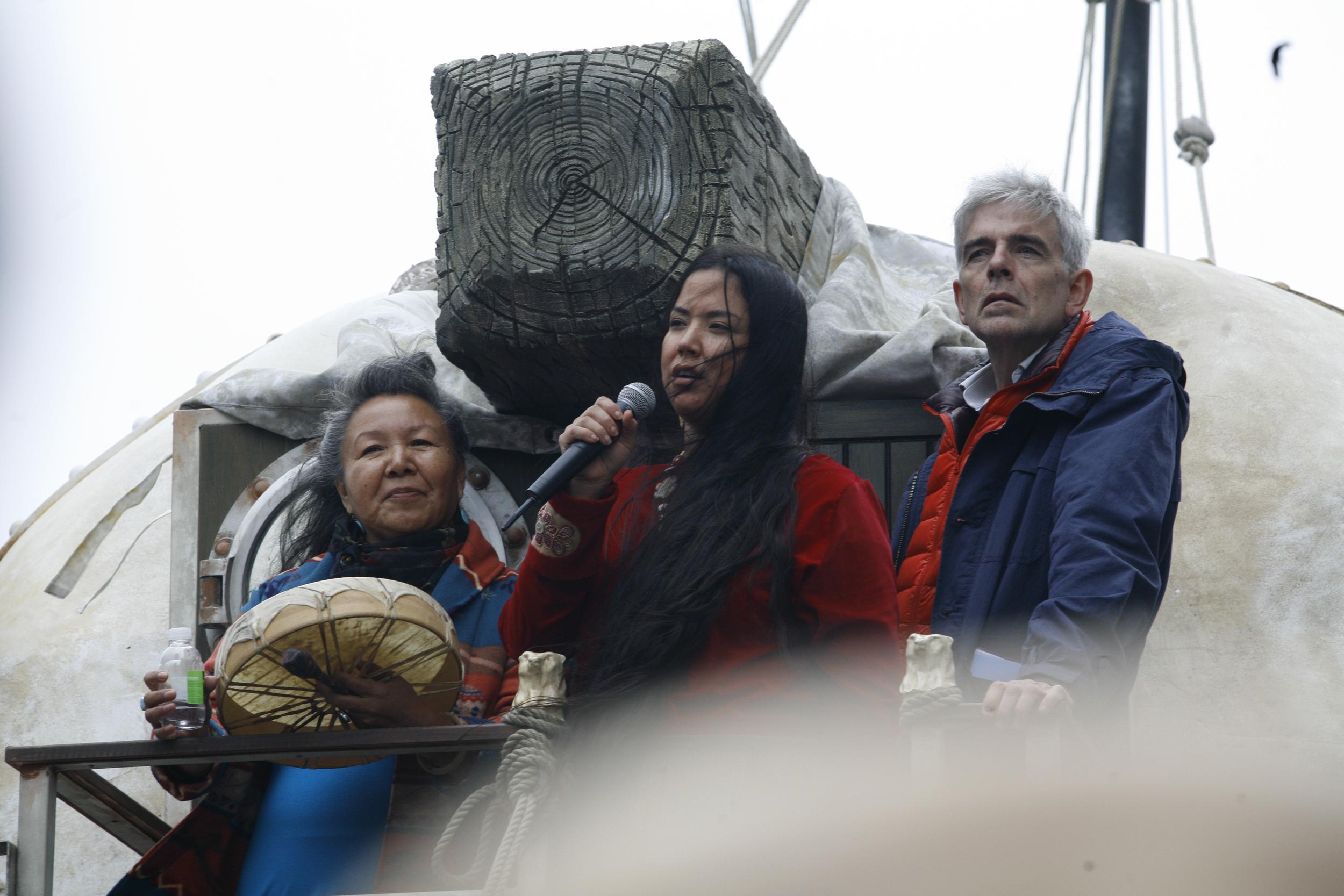 Three activists stand on a stage in cold weather gear. One speaks into a microphone.