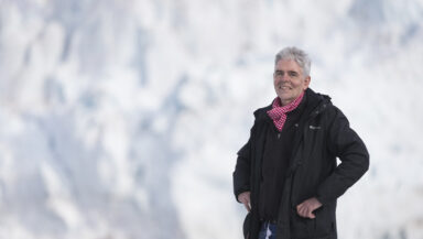John Sauven, dressed in cold weather gear, smiles in a casual pose, standing in front of a white wall of ice.