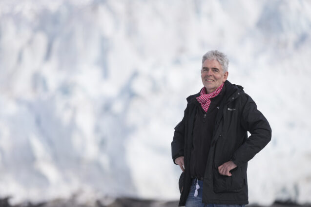 John Sauven, dressed in cold weather gear, smiles in a casual pose, standing in front of a white wall of ice.