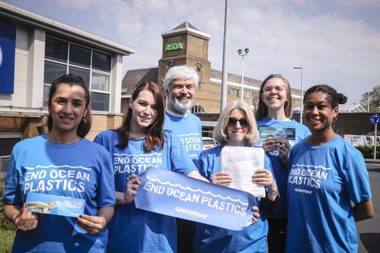 A group of six Greenpeace volunteers of different ages and ethnicities stand in the sunshine outside an ASDA supermarket. Smiling into the camera, they wear blue tshirts with 'End ocean plastics' slogans, and hold up campaign banners.