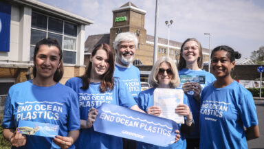 A group of six Greenpeace volunteers of different ages and ethnicities stand in the sunshine outside an ASDA supermarket. Smiling into the camera, they wear blue tshirts with 'End ocean plastics' slogans, and hold up campaign banners.
