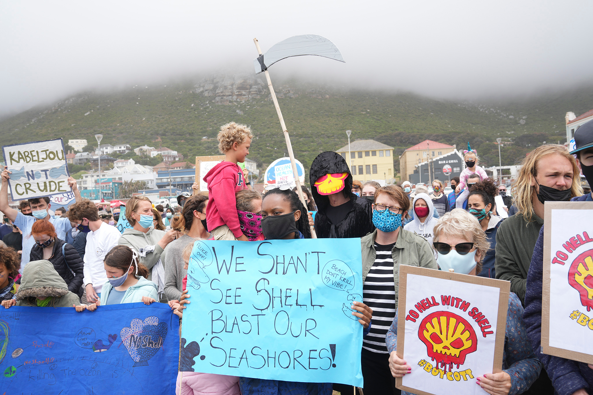 A crowd of protestors hold up signs criticising Shell. One at the front reads 'We shan't see Shell blast our seashore'. Another protestor is dressed as the Grim Reaper, with a black cape and Scythe, plus a Shell logo over their face.