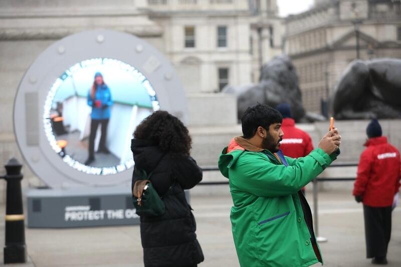 A passerby takes a selfie in front of a large circular outdoor screen set up on a plinth amongst the grand stone buildings of Trafalgar Square. The screen shows someone standing on the deck of a ship in a snowy landscape, speaking to the camera.