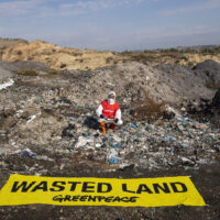 An activist in protective overalls, goggles and a mask kneels in front of a large pile of ash and burned plastic. Laid out in the foreground is a large banner reading 'Wasted land'