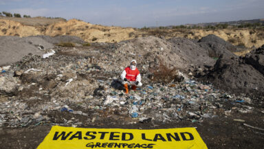 An activist in protective overalls, goggles and a mask kneels in front of a large pile of ash and burned plastic. Laid out in the foreground is a large banner reading 'Wasted land'