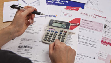 A view of a range of utility bills and a calculator spread out on a table, seen over the shoulder of someone holding a pen