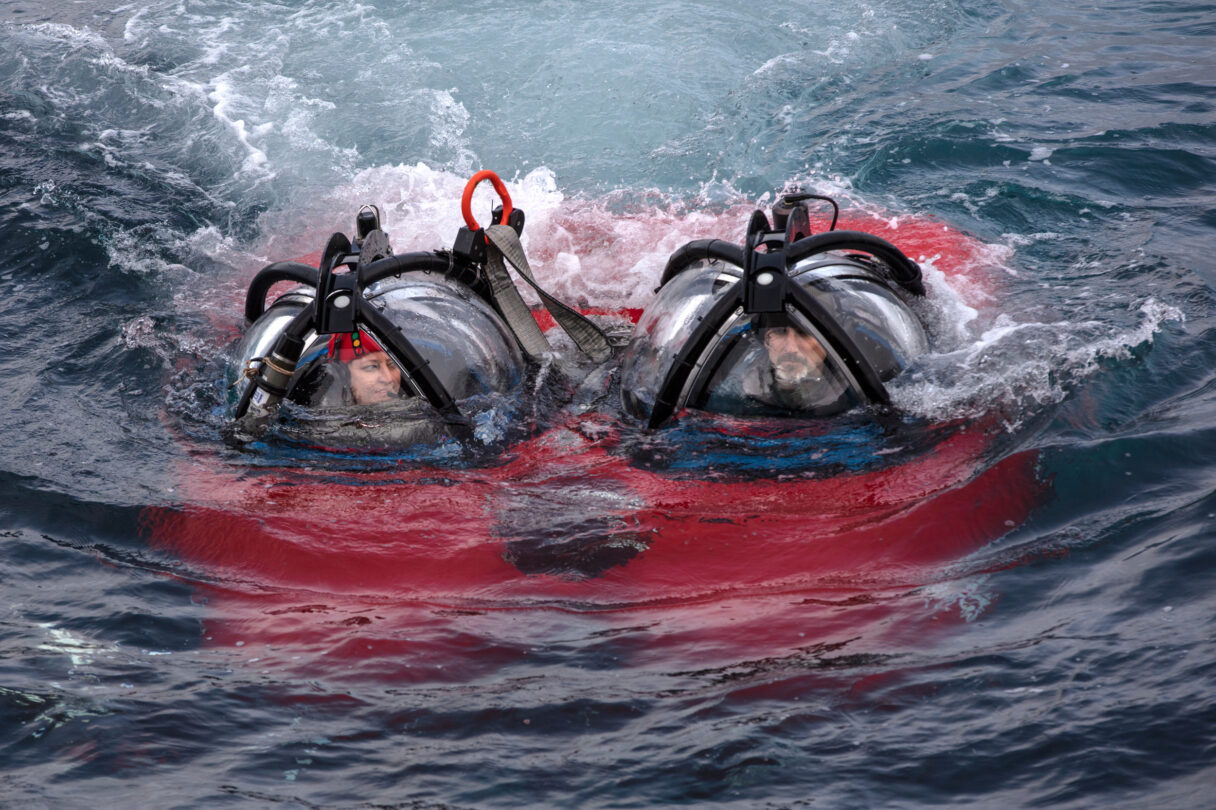 A small red submarine emerges from dark water, clear domes on top reveal two people inside