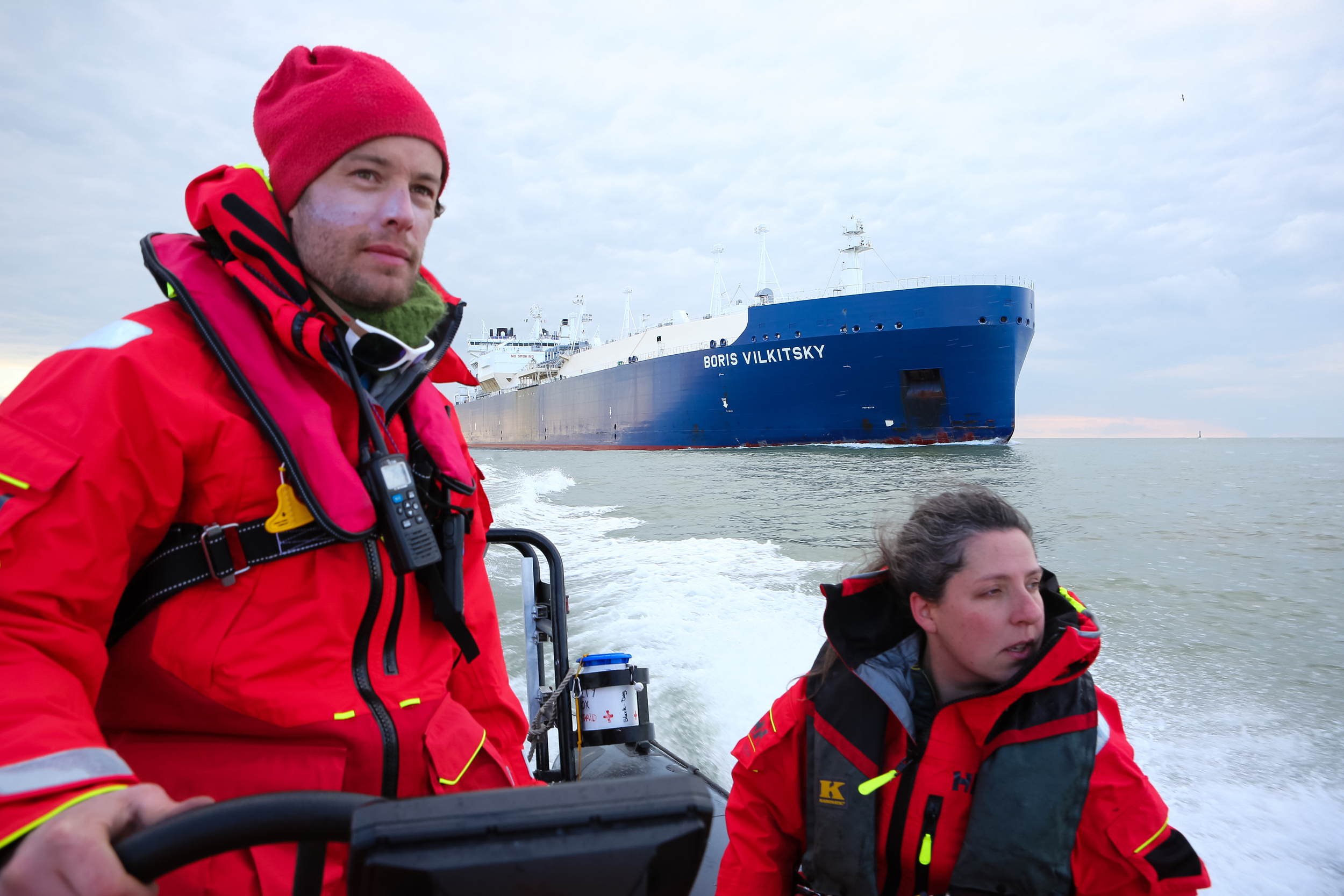 Two people in a rigid-hulled inflatable boat travelling in open water, a large oil tanker in the background