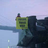 A Greenpeace activist in climbing gear hangs from the edge of a huge commercial ship dock, holding a banner that says 'Fossil fuels war'. In the background, there's a giant concrete suspension bridge, and the milky pink sky of the early dawn.