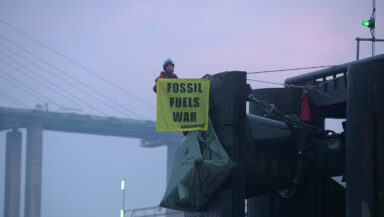 A Greenpeace activist in climbing gear hangs from the edge of a huge commercial ship dock, holding a banner that says 'Fossil fuels war'. In the background, there's a giant concrete suspension bridge, and the milky pink sky of the early dawn.