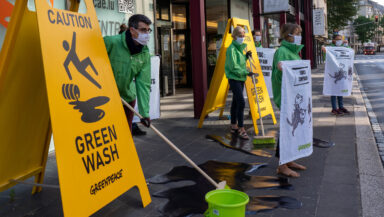 Activists dressed in green boiler suits mop the floor outside Luxembourg’s pension fund. They place yellow hazard A-board floor signs nearby that read “Caution green wash” with a symbol of someone slipping over.
