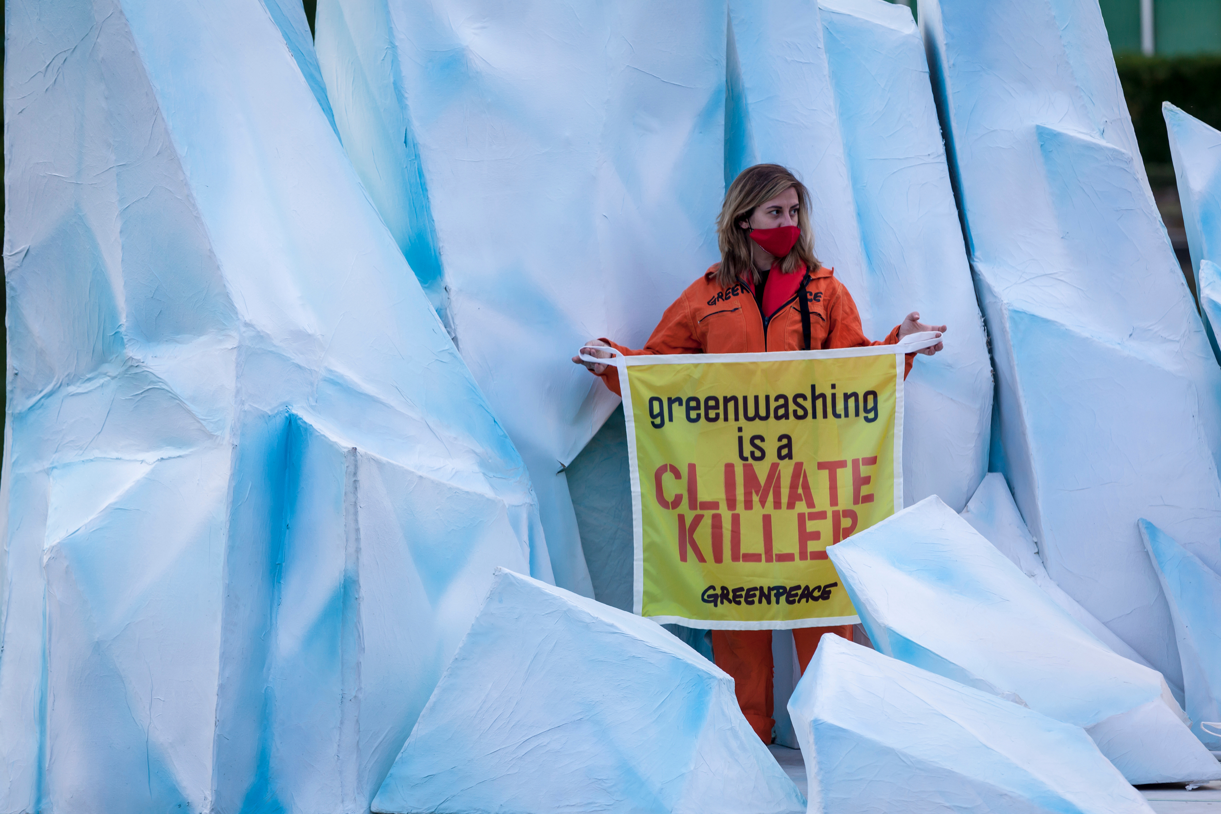 Activist stands holding a sign that reads “Greenwashing is a CLIMATE KILLER”. They stand on a floating reproduction of a melting iceberg.