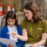 A volunteer wearing a Greenpeace t-shirt sits on a bench outside with a primary school pupil. The volunteer is speaking and pointing to a sheet of paper held by the pupil.