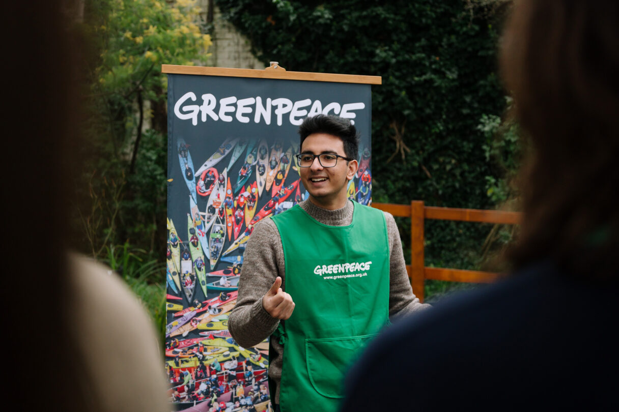 A volunteer wearing a Greenpeace branded tabard stands in front of a colourful Greenpeace banner speaking happily and confidently to an unseen audience.