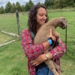 Sandra is standing in a field smiling and holding a cute baby goat