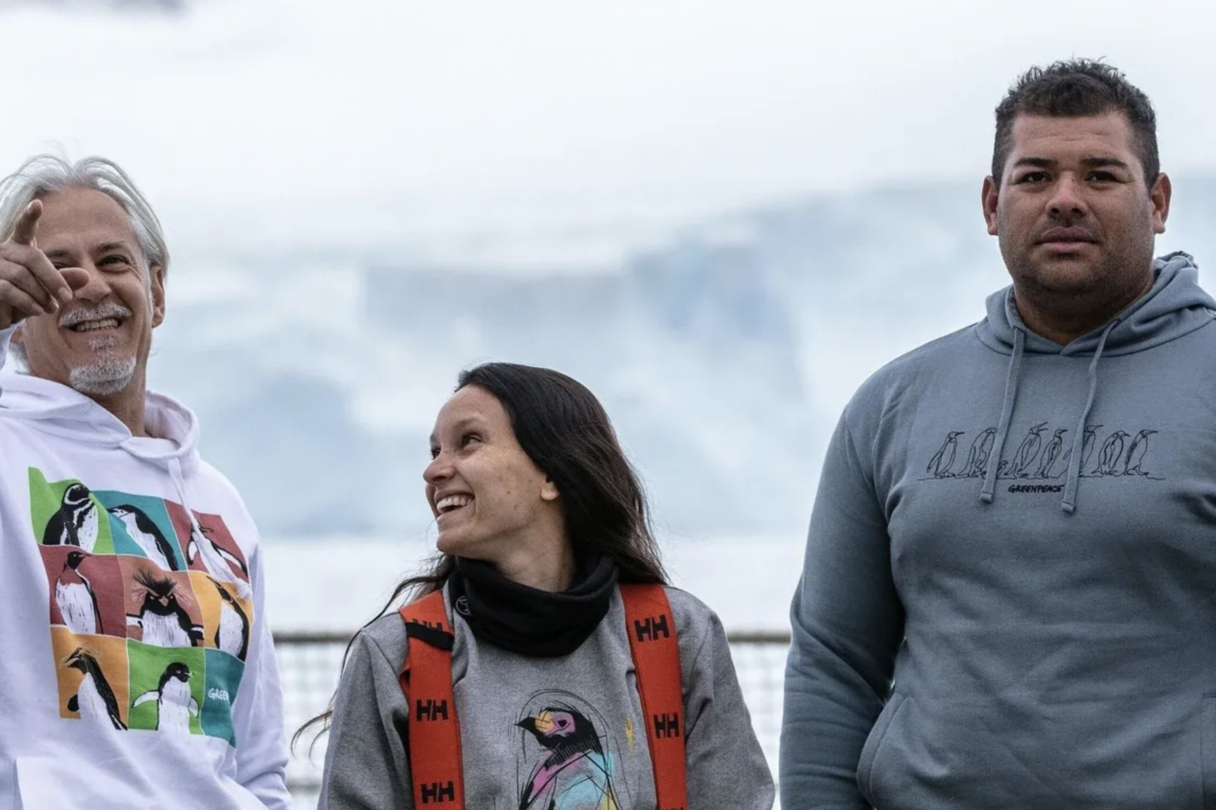 Three Greenpeace crew members wearing branded sweaters with penguin designs. They stand on the deck of a ship in a relaxed, informal pose, with a glacier in the background.
