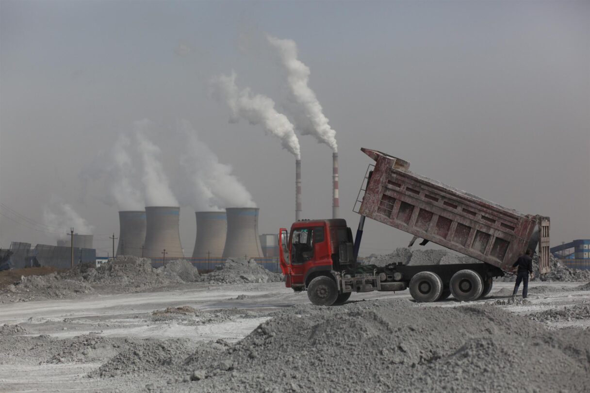 A truck carrying coal ash in a desolate and bleak landscape. There are coal chimneys in the distance with smoke going out into the atmosphere.