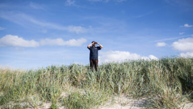 Person looking through binoculars. They stand in green grass by a beach with a blue sky above them.