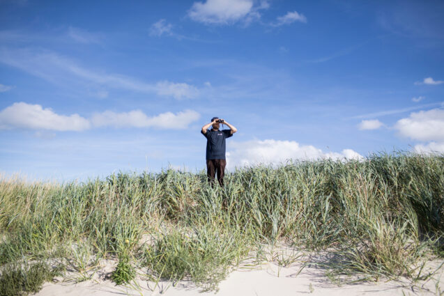 Person looking through binoculars. They stand in green grass by a beach with a blue sky above them.