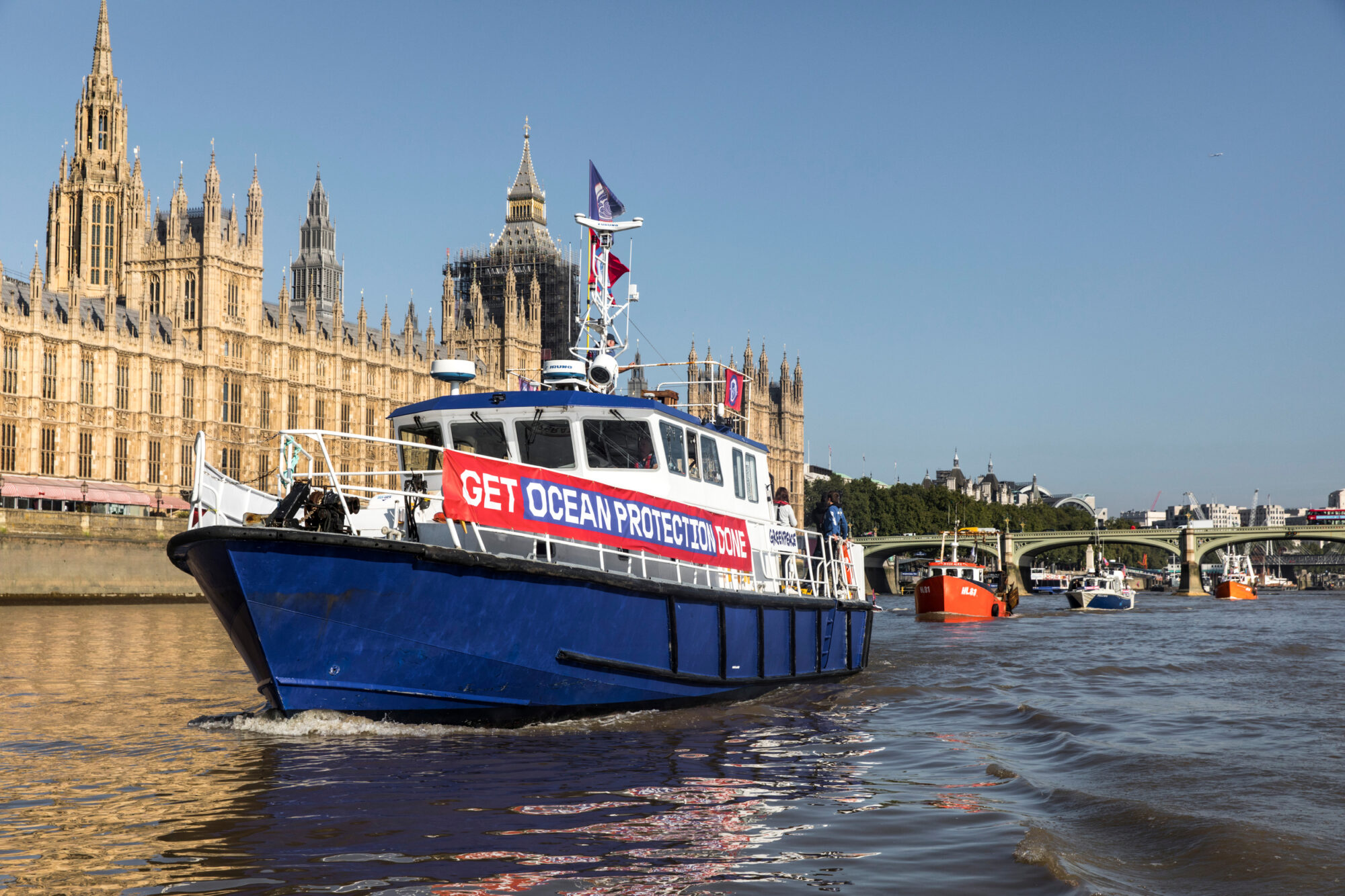 A fishing boat on the river Thames outside the Houses of Parliament. A sign on the side reads "Get ocean protection done"