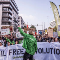 A crowd of protesters stands in a city street holding banners and placards, including one saying 'Fossil free revolution'. At the front, a person wearing a Greenpeace jacket raises a fist to rally the crowd.
