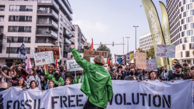 A crowd of protesters stands in a city street holding banners and placards, including one saying 'Fossil free revolution'. At the front, a person wearing a Greenpeace jacket raises a fist to rally the crowd.