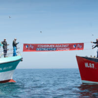 French and English fishermen in the middle of the water. They hold a sign between the two boats that reads "Fishermen against destructive fishing"
