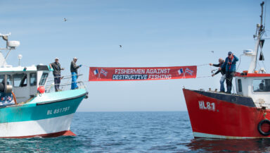 French and English fishermen in the middle of the water. They hold a sign between the two boats that reads "Fishermen against destructive fishing"