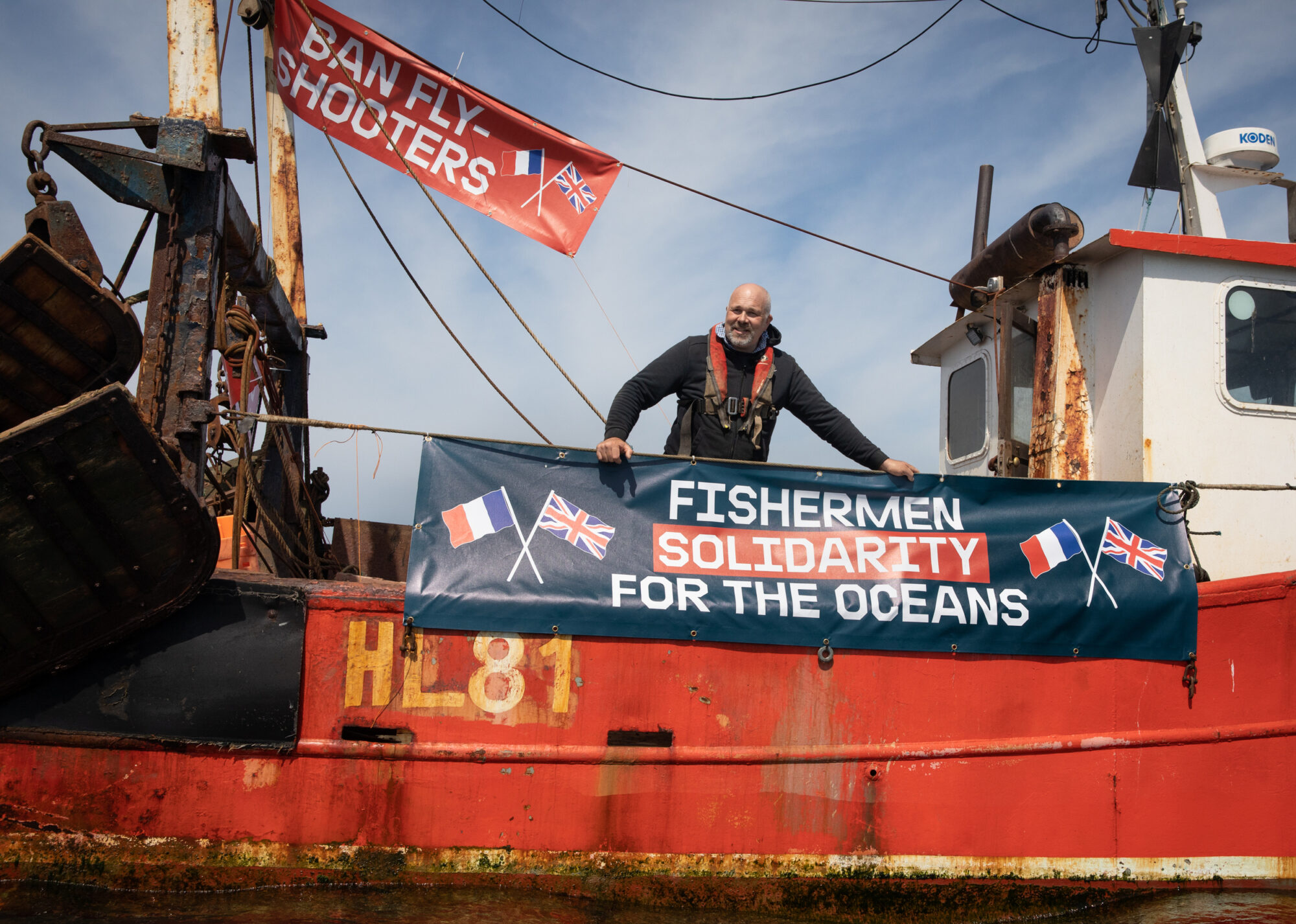 Fisherman stands on deck of a boat with a banner saying Fishermen solidarity for the oceans with crossed french and uk flags