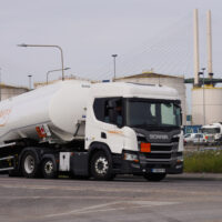 A tanker lorry with the Sainsburys logo on the side pulls away from a terminal complex. White industrial silos are visible in the background.