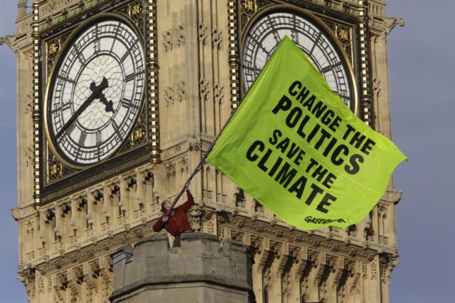An activist stands on top of a stone tower waving a huge flag with the slogan "change the politics save the climate". Behind him, the iconic Big Ben clock looms just out of focus.