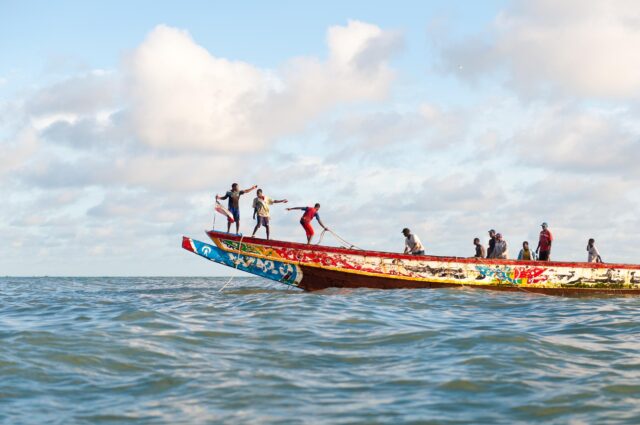 Senegalese fishermen stand on the bow of a wooden fishing boat painted in a colourful traditional design.