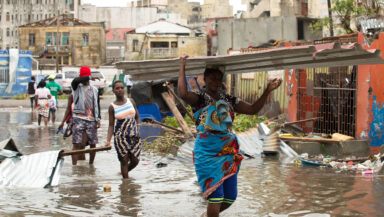 Black people wade along a flooded city street, knee-deep in dirty water and surrounded by debris. In the foreground, one person carries a large piece of corrugated metal on her head.