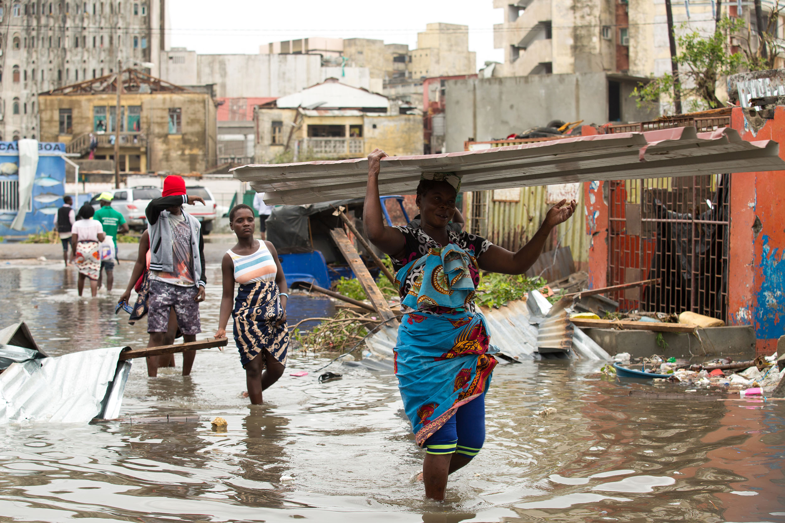Black people wade along a flooded city street, knee-deep in dirty water and surrounded by debris. In the foreground, one person carries a large piece of corrugated metal on her head.