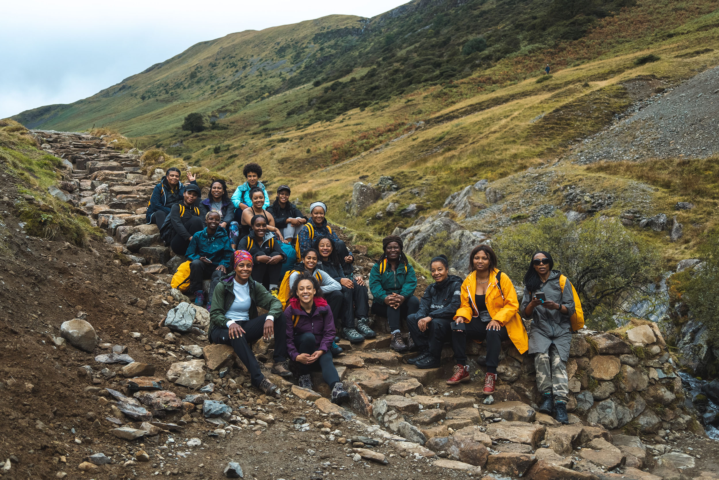 A group of around 20 Black and brown people in hiking gear smile for a photo on a rocky hillside.
