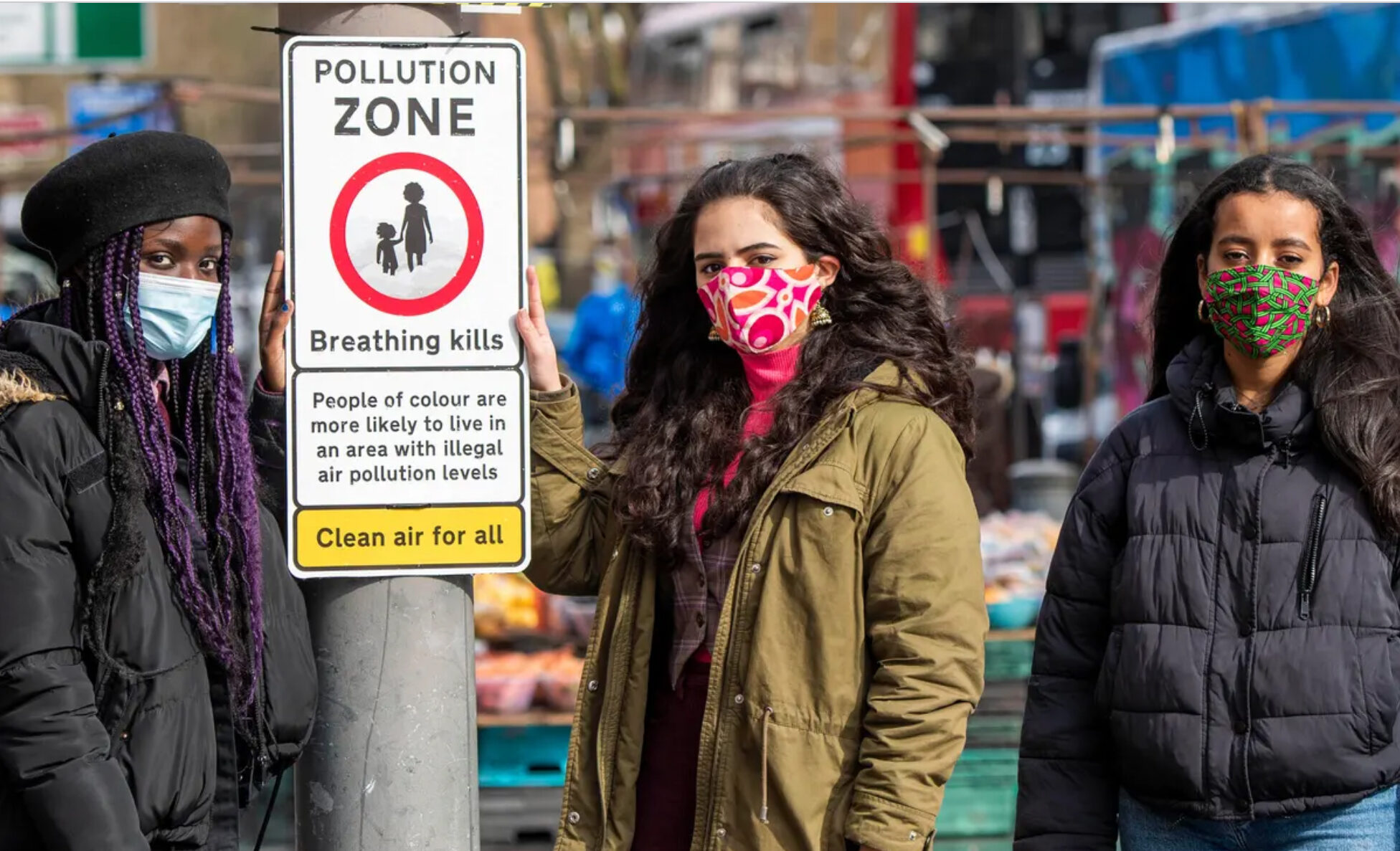 Three young people wearing winter jackets and facemasks stand on a city street next to a sign board mounted on a pole. Designed in the style of an official road sign, the board shows a parent and child in a red circle. Text warns about dangerous air pollution in the area.
