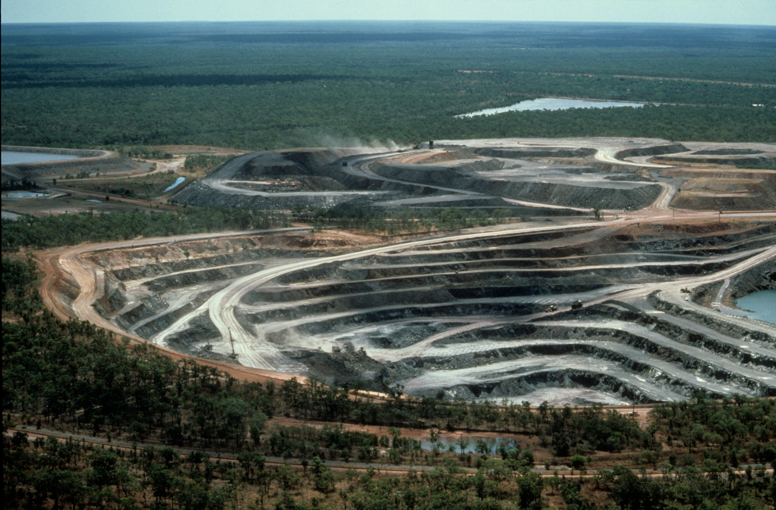 Aerial view of an open-cast mine in the middle of a forest. A huge, wide hole has been cut into the earth, sloping down in a series of flat terraces.