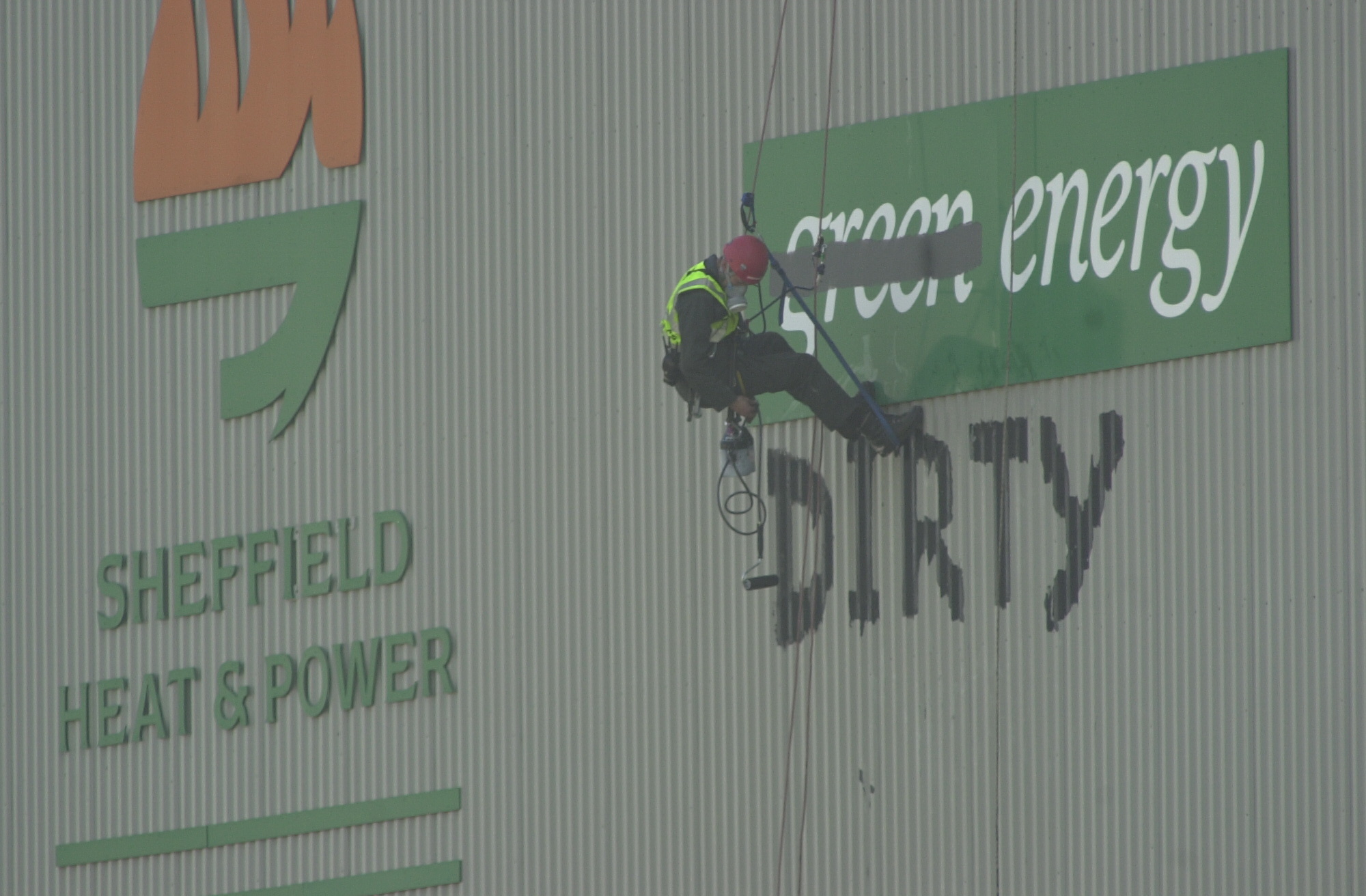 Climber on side of building painting over sign "green energy" to read "dirty". Other sign says "Sheffield Heat and Power".