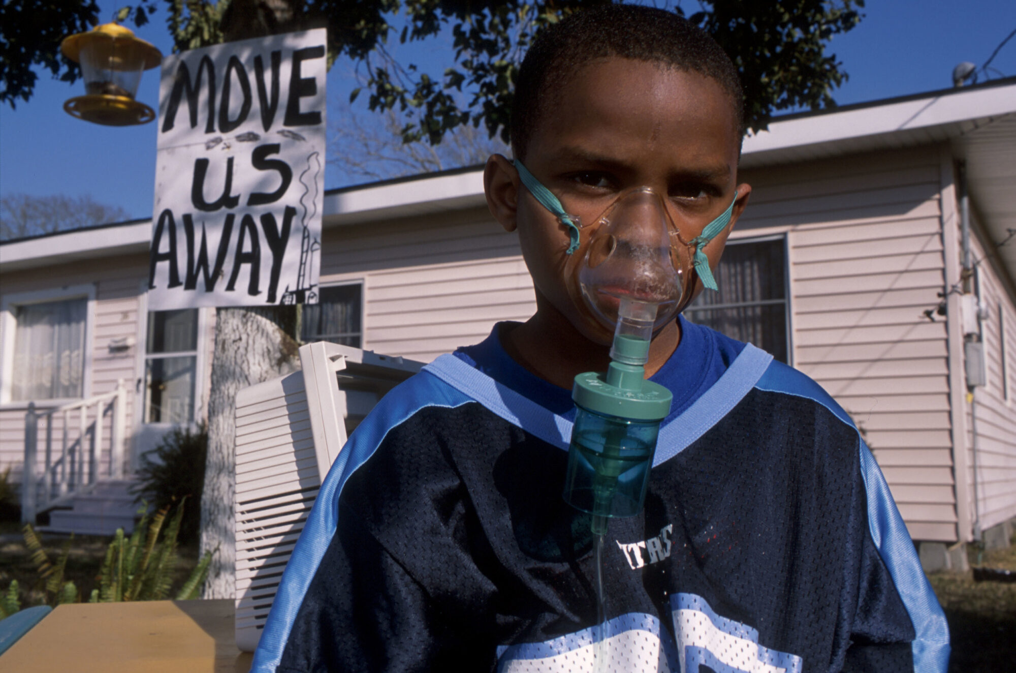 A Black child wearing an oxygen mask looks into the camera. Behind him is a white clapboard house with a hand-painted sign reading 'Move us away'.