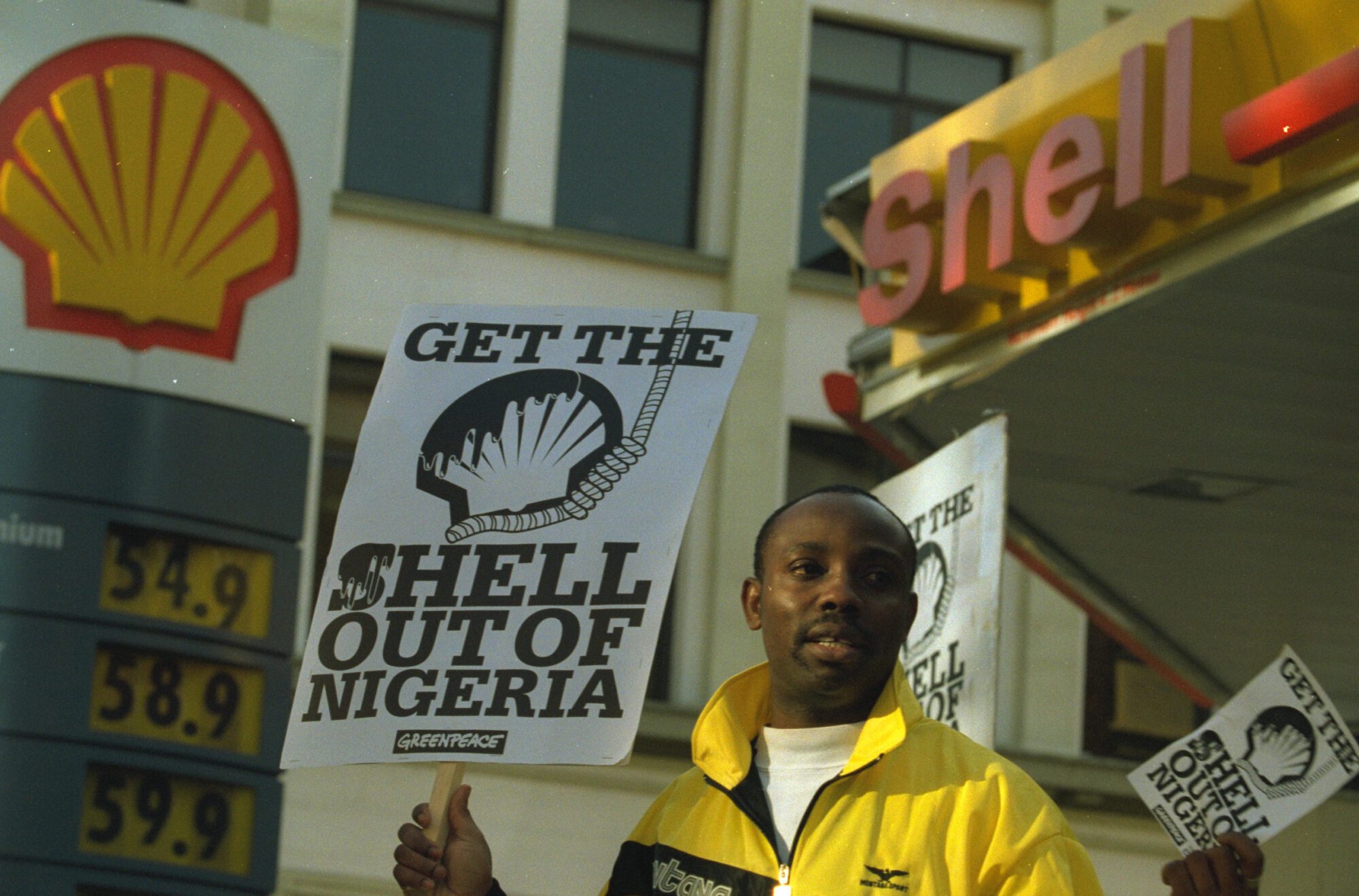 A Black man stands in front of a Shell petrol station holding a placard that reads 'Get the Shell out of Nigeria'. More identical placards are visible over his shoulder.