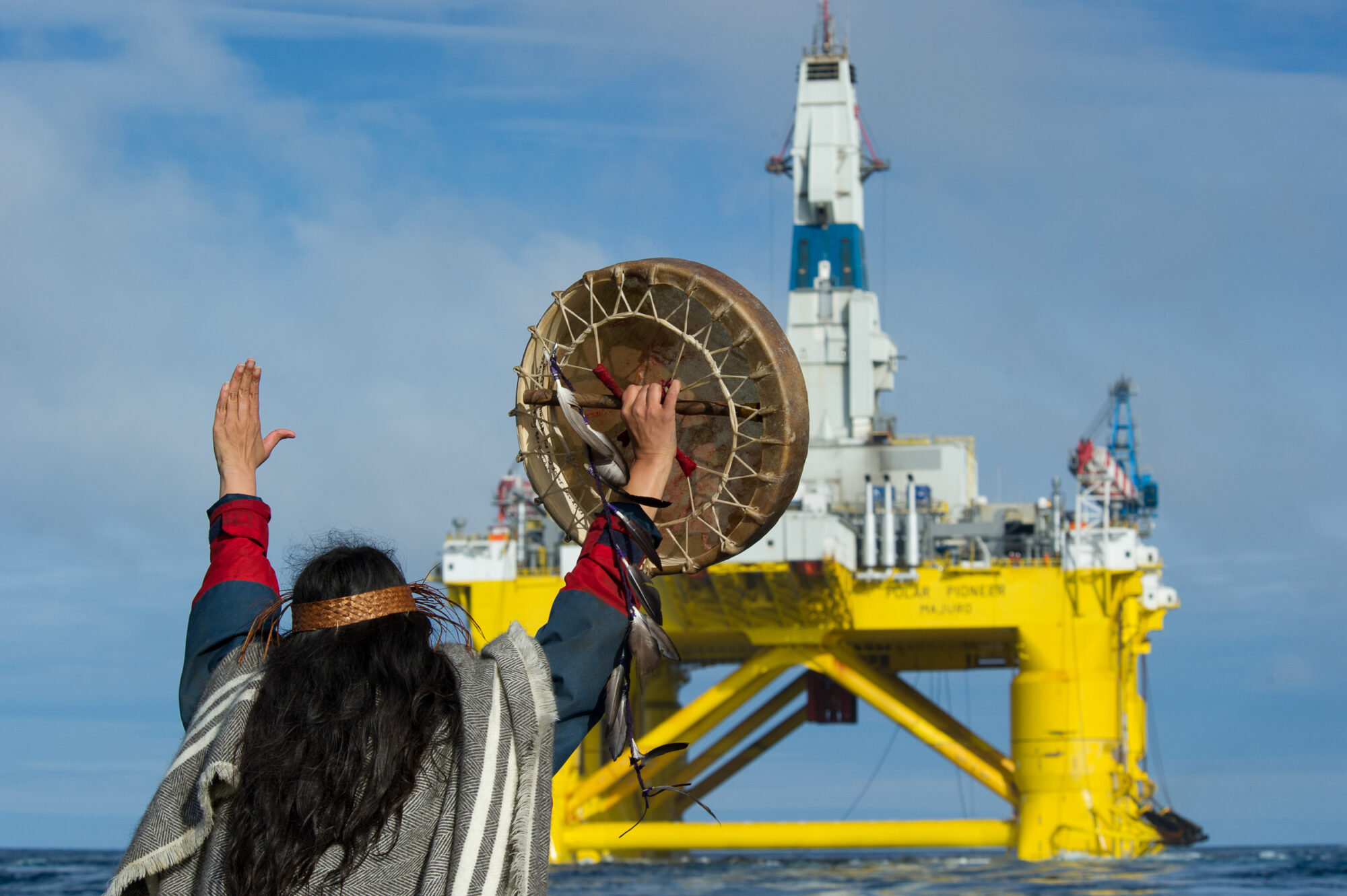A person in traditional Indigenous Musqueam dress defiantly raises their hands towards a floating oil platform close to the shore. In one hand they're holding a drum.