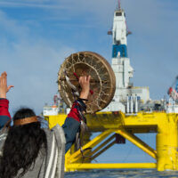 A person in traditional Indigenous Musqueam dress defiantly raises their hands towards a floating oil platform close to the shore. In one hand they're holding a drum.