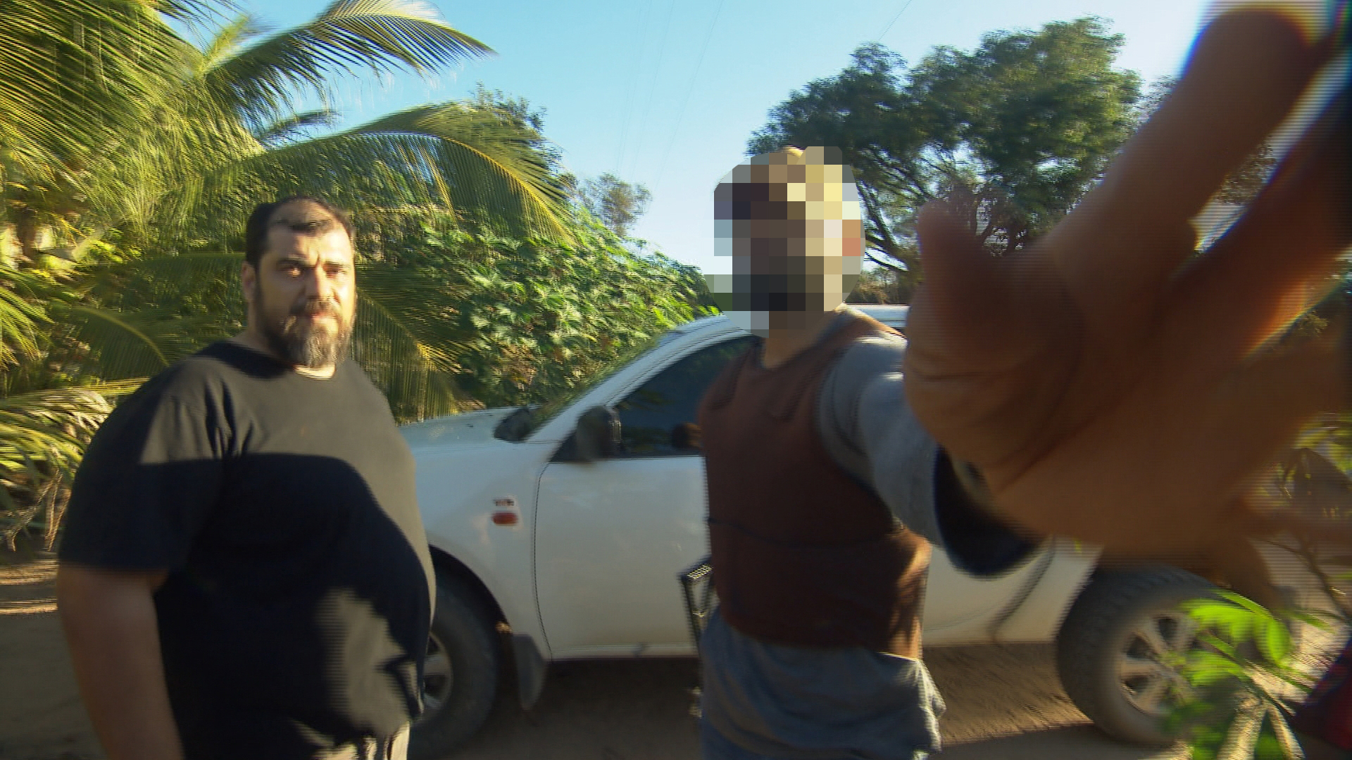 Two people stand in front of a car, with tropical foliage in the background. One, with their face pixelated, reaches out to obstruct the camera lens.