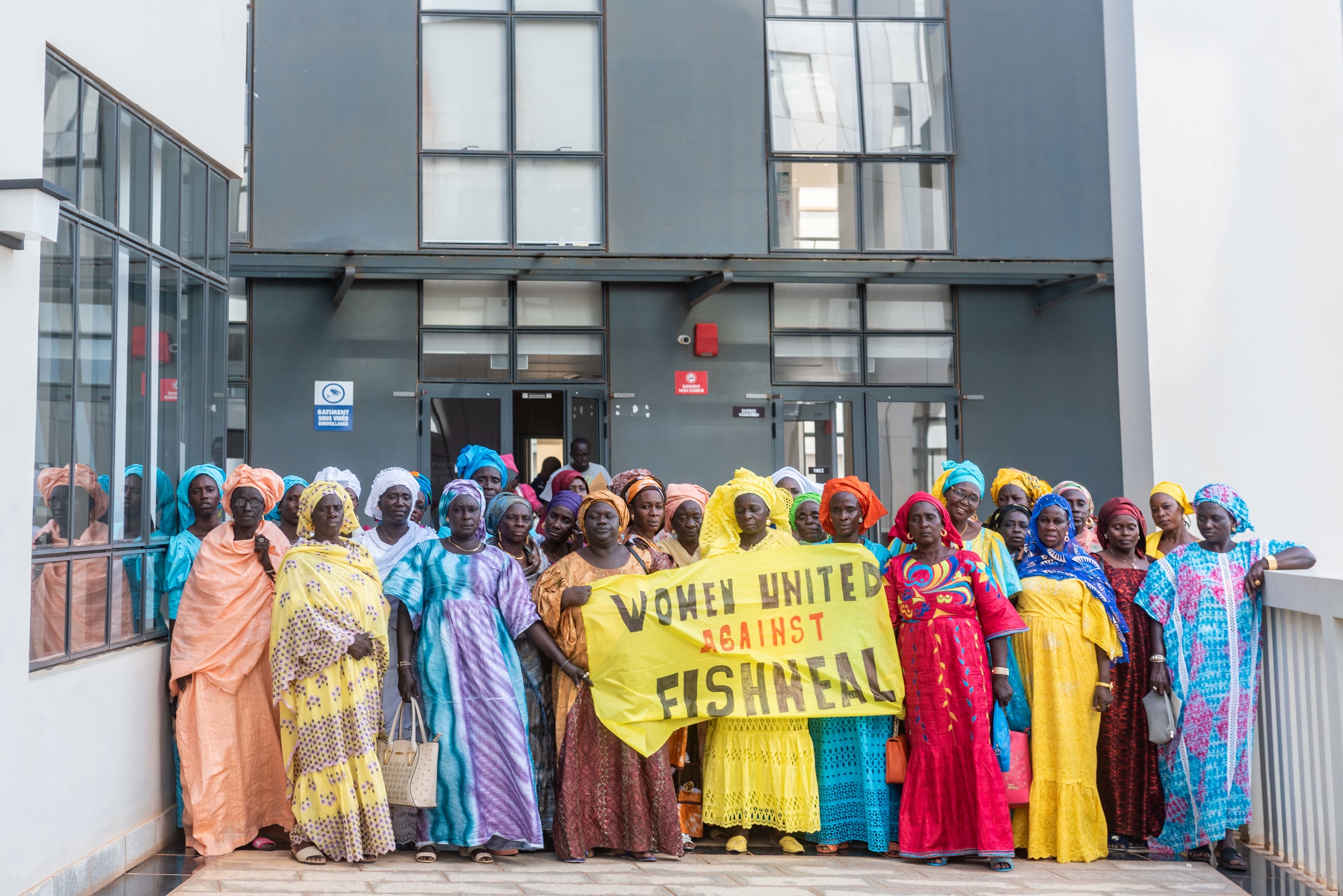 Around 30 Black women in long, colourful dresses and matching headscarves stand close together facing the camera. At the front of the group, one holds a hand-painted banner reading 'Women united against fishmeal'.