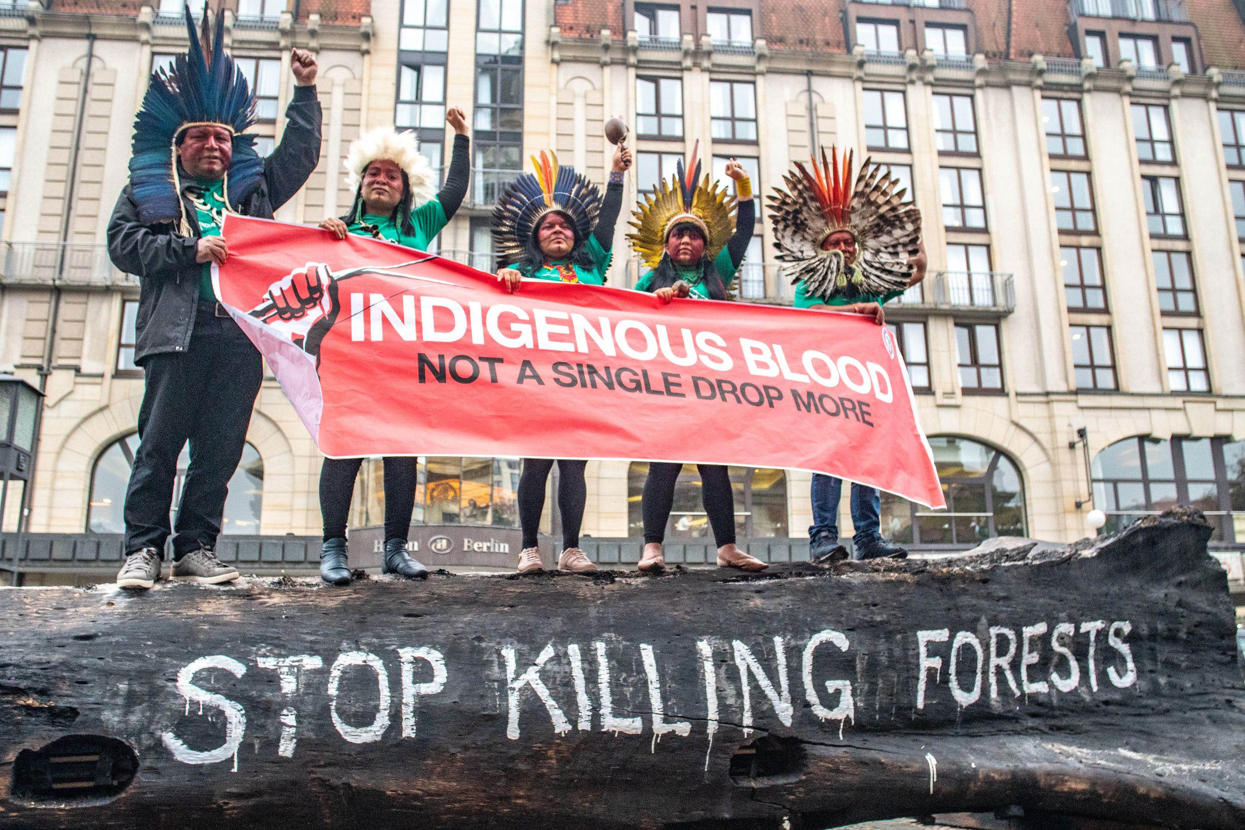 Five people hold a large banner reading 'Indigenous blood: not a single drop more'. Fists raised defiantly in the air, they're wearing feathered headdresses typical of the Indigenous Peoples of the Amazon.