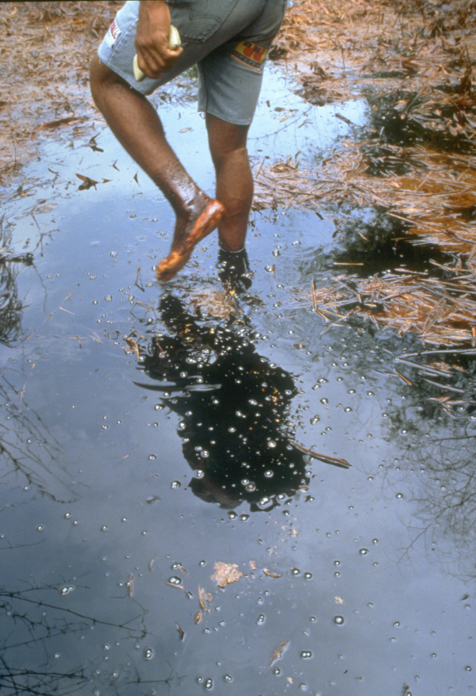 Legs and feet of a Black person walking barefoot through shallow water thickly coated with oil.