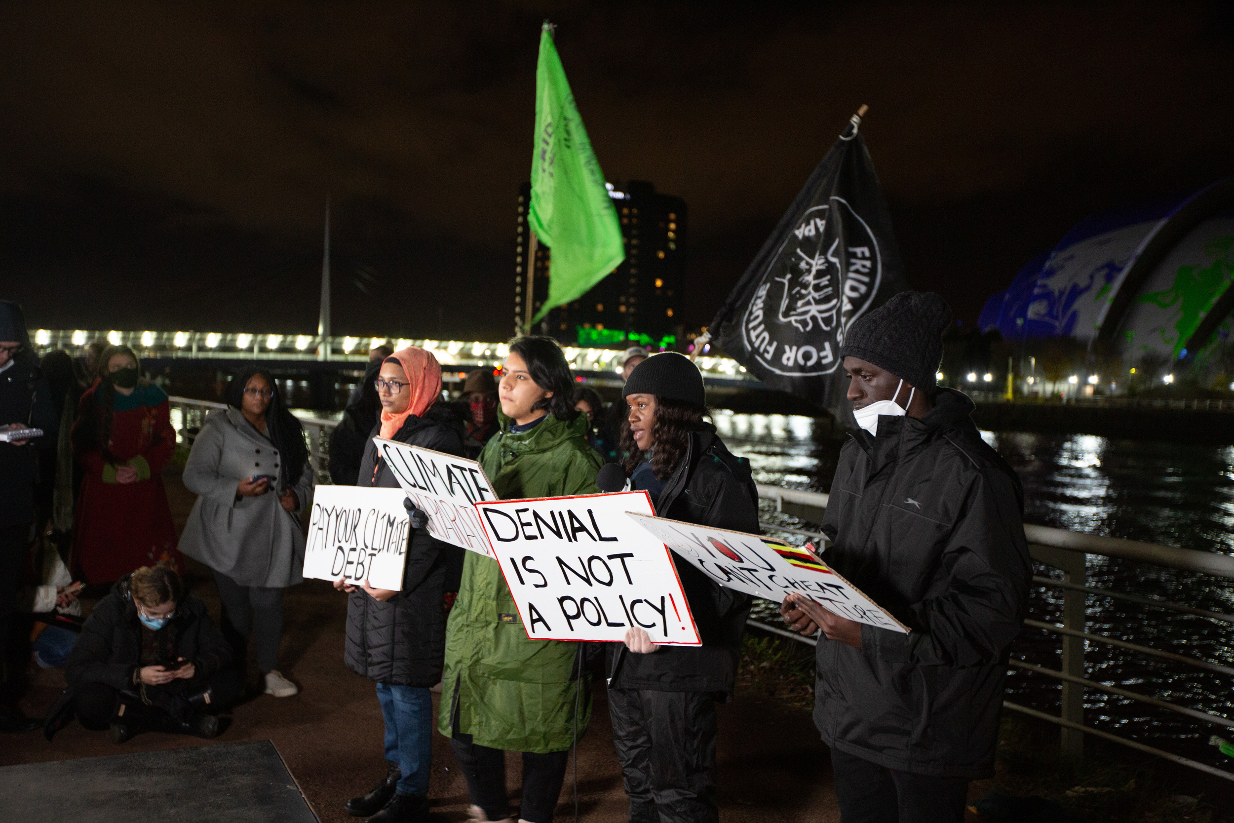 Four young people of colour stand with their backs to the water in a city dockside at night time. Two are Black, and two have light brown skin. One wears a headscarf. They hold placards with slogans including 'Denial is not a policy' and 'Pay your climate debt'.
