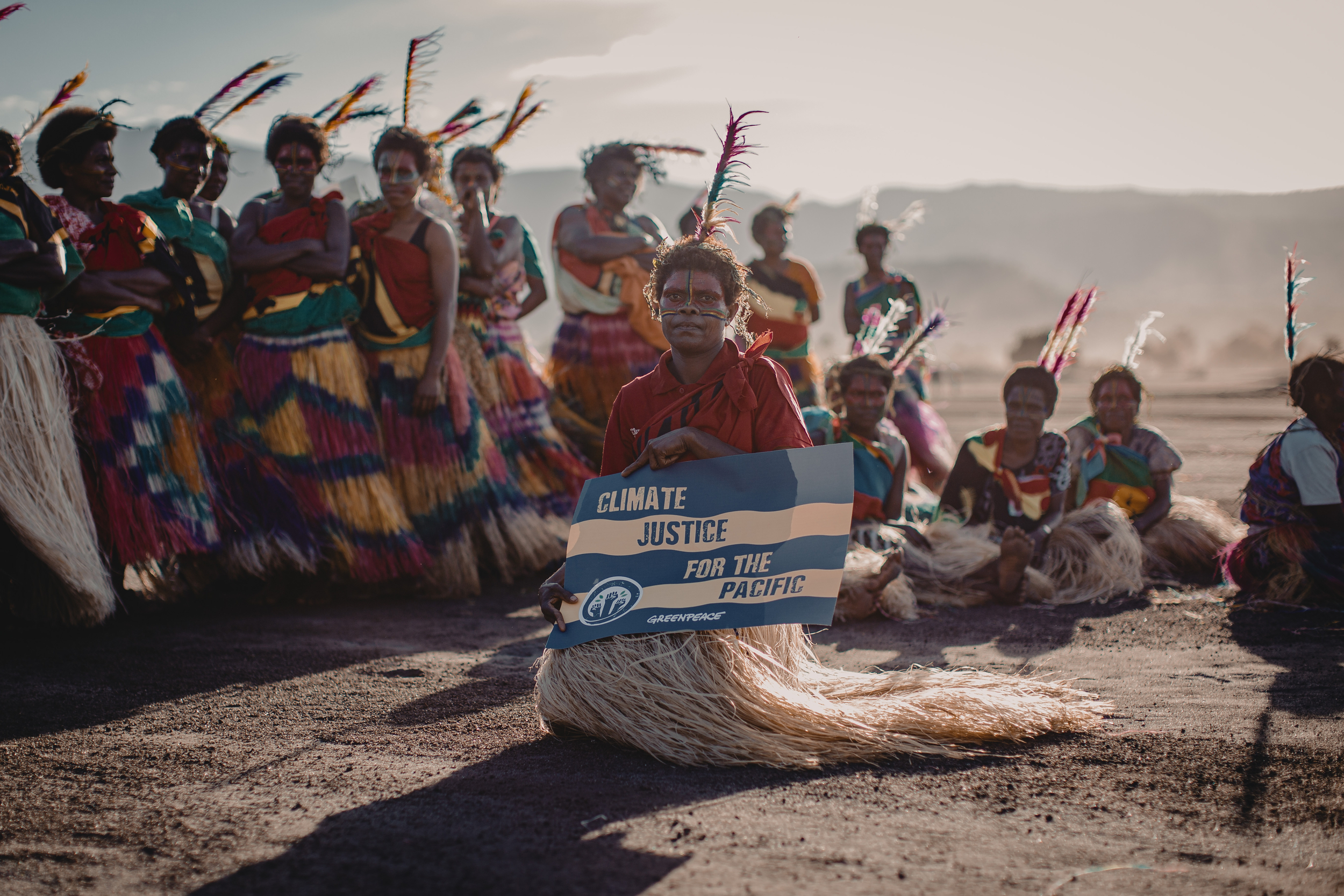 15-or-so brown-skinned women are gathered on a bright, windy beach or desert landscape. They wear colourful grass skirts, red, green and gold shawls, and decorative headdresses. At the front, one holds a Greenpeace branded sign reading 'Climate justice for the Pacific'.