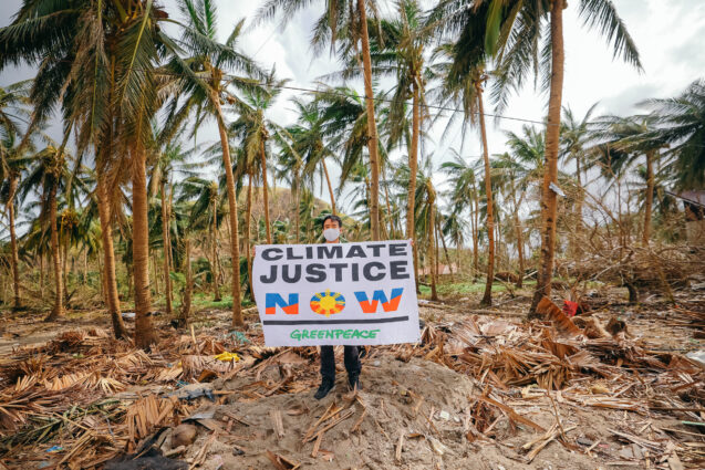 Person holding a sign that reads “Climate justice now”. There are broken palm trees behind them and debris covering the floor they stand on.