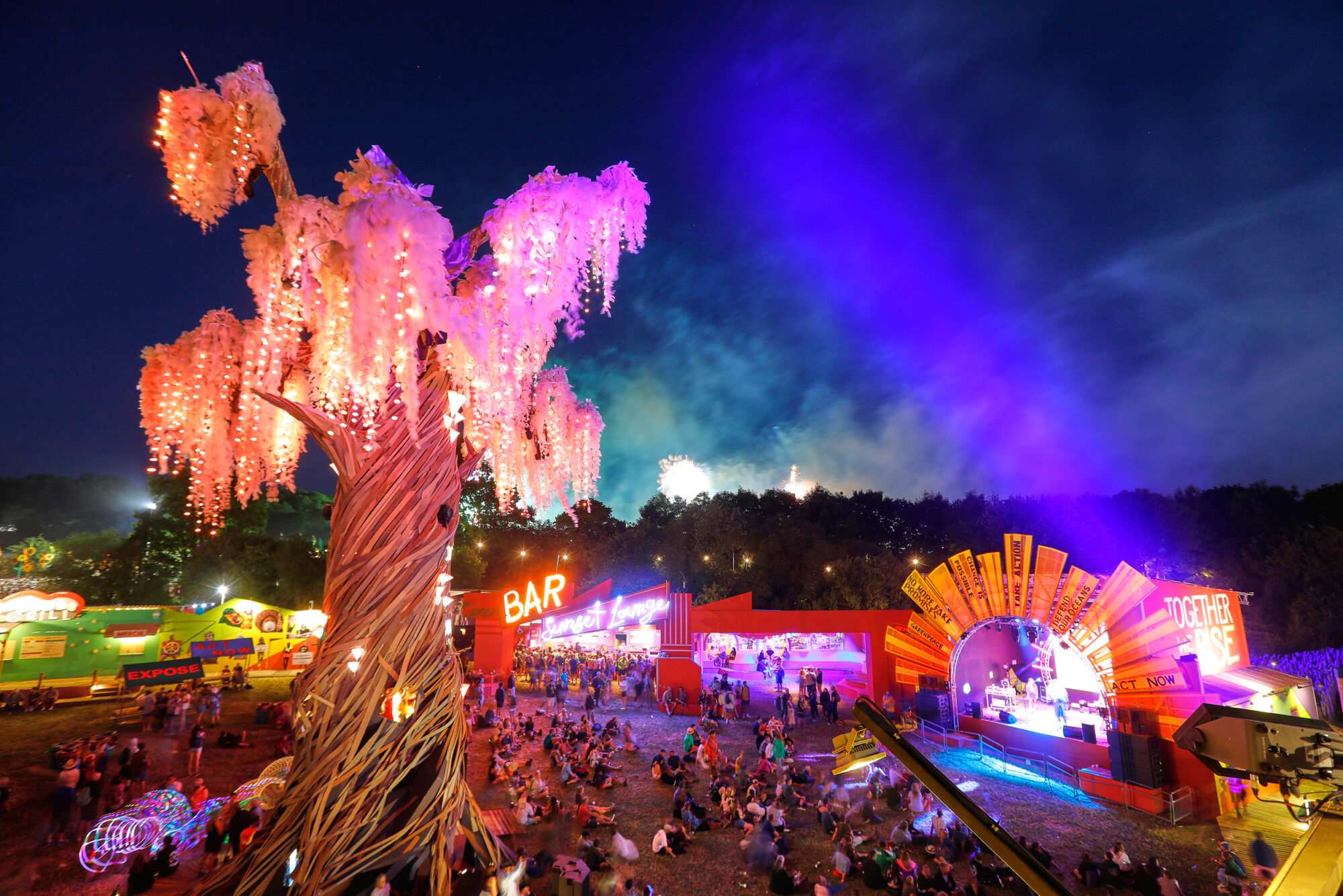 A giant artificial tree lit up with fairy lights overlooks a huge festival crowd at night.